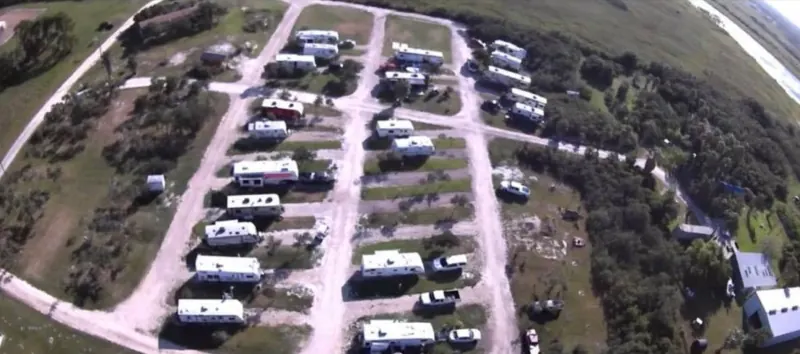 Aerial view of Bay Breeze RV Park showing rows of RV sites along gravel drives, with grass, trees and open ground around them