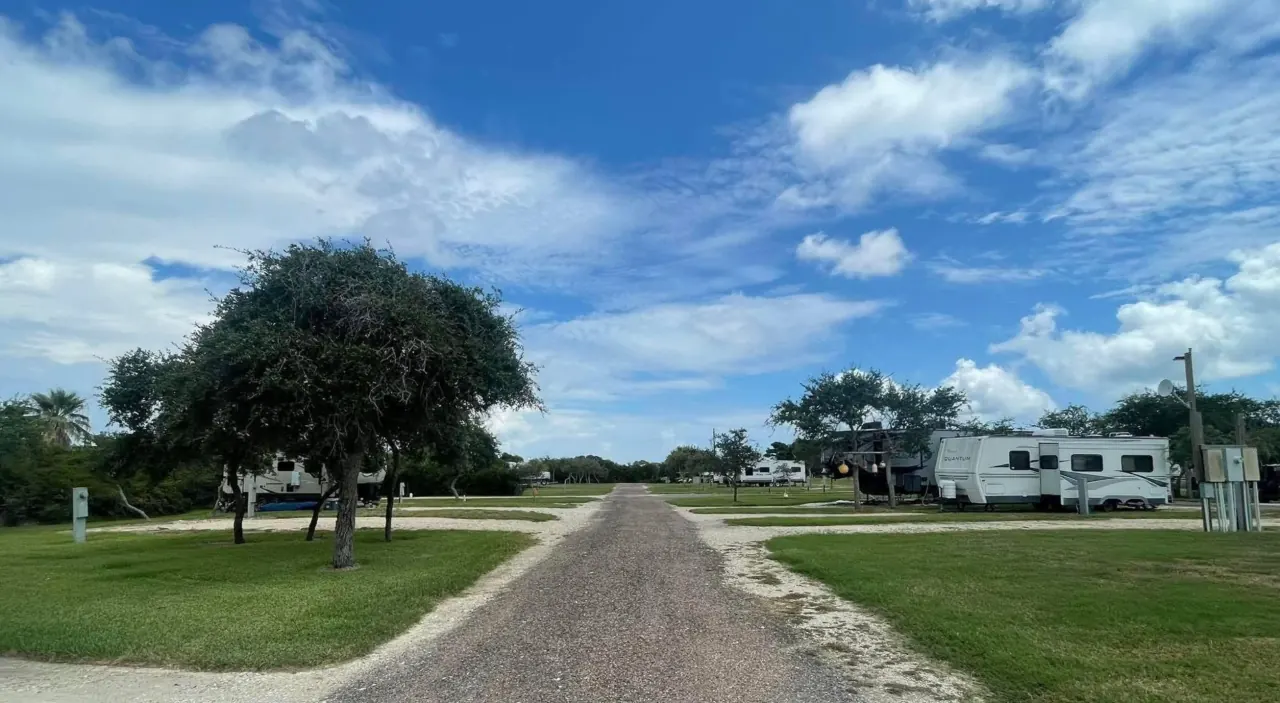 Gravel entrance road at Bay Breeze RV Park with RV sites on both sides, a live oak shading the left verge and open blue sky ahead